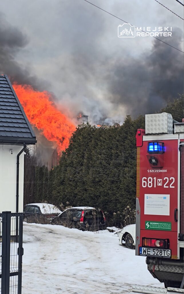 Płonący budynek otoczony gęstym dymem, w tle widoczny wóz strażacki z włączonymi sygnalizatorami.