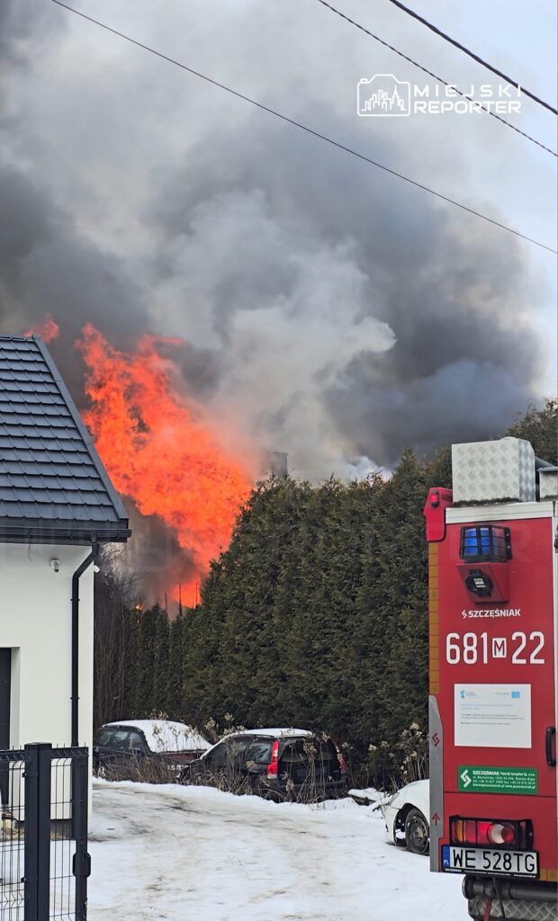 Płomienie i gęsty dym unoszą się nad drzewami, obok stoi wóz strażacki na śnieżnej drodze.