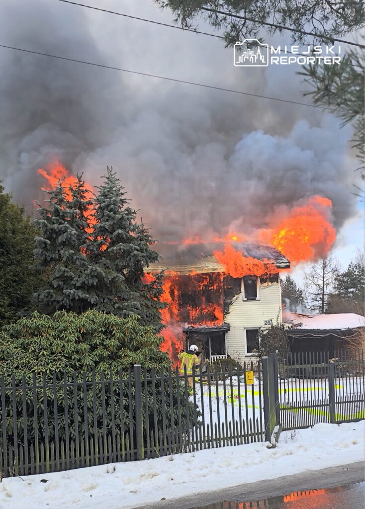Dom w płomieniach, ogień wydobywający się z dachu, strażak w żółtej kamizelce gaszący pożar w zimowej scenerii.