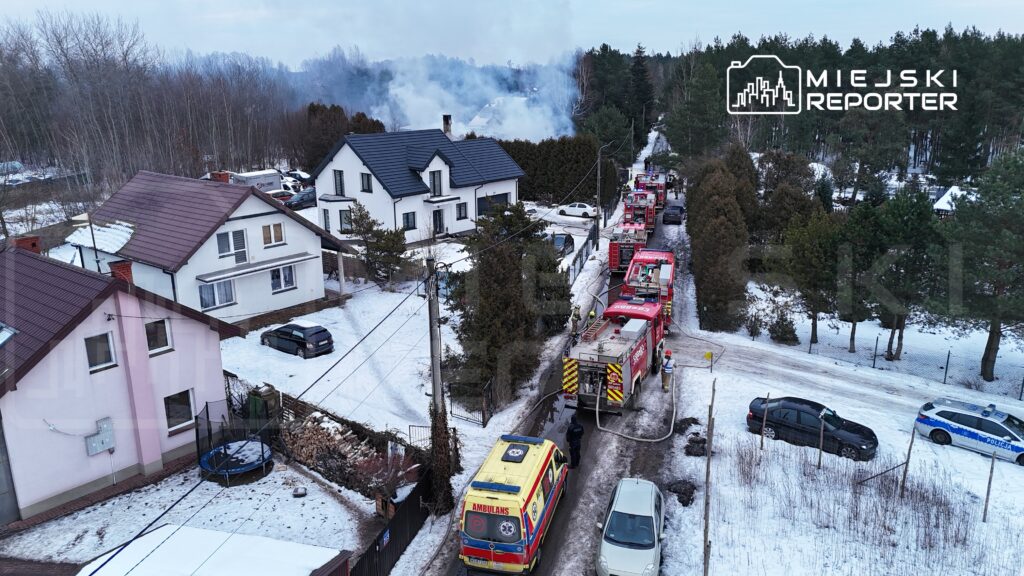 W pobliżu domów widać pojazdy straży pożarnej oraz ambulans, a w tle unosi się dym z pożaru.