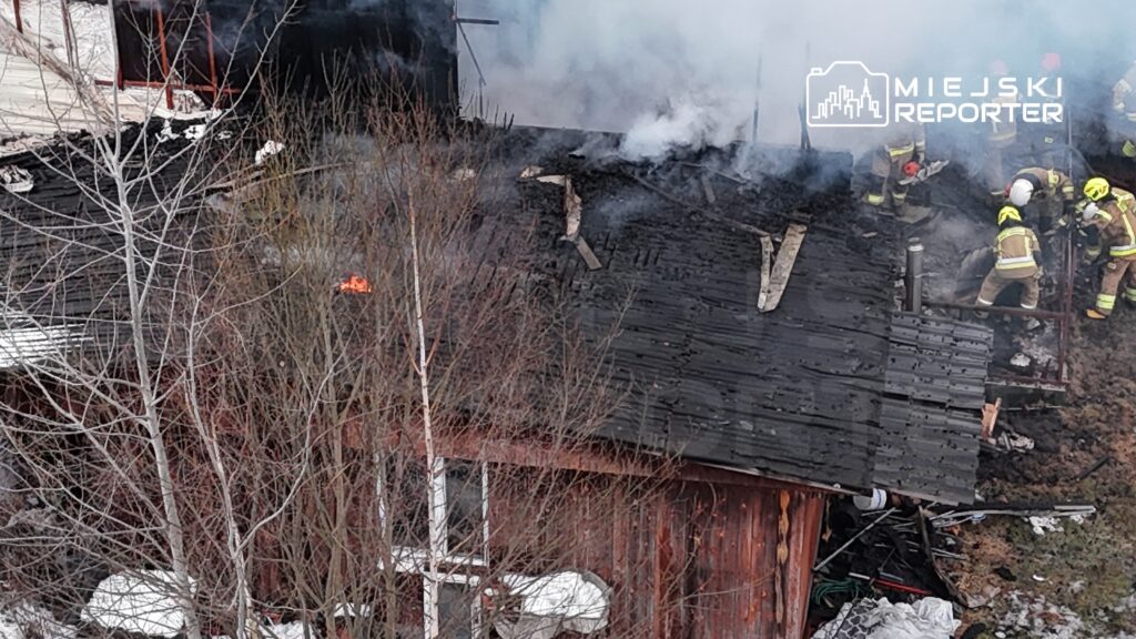 Strażacy gaszą pożar dachu drewnianego budynku, z którego wydobywają się kłęby dymu. W tle widać uszkodzone elementy konstrukcji.