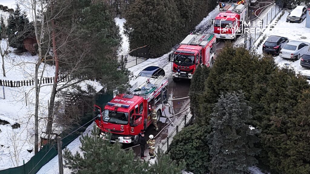 Dwa samochody strażackie zaparkowane na śnieżnej drodze, a strażacy rozmawiają z mężczyzną w czarnym płaszczu.
