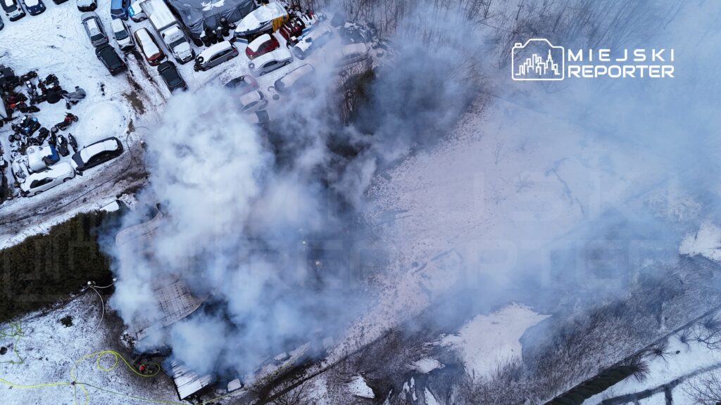 Dym unoszący się z płonącego obiektu w zalesionym terenie, otoczonym śniegiem i zaparkowanymi samochodami.
