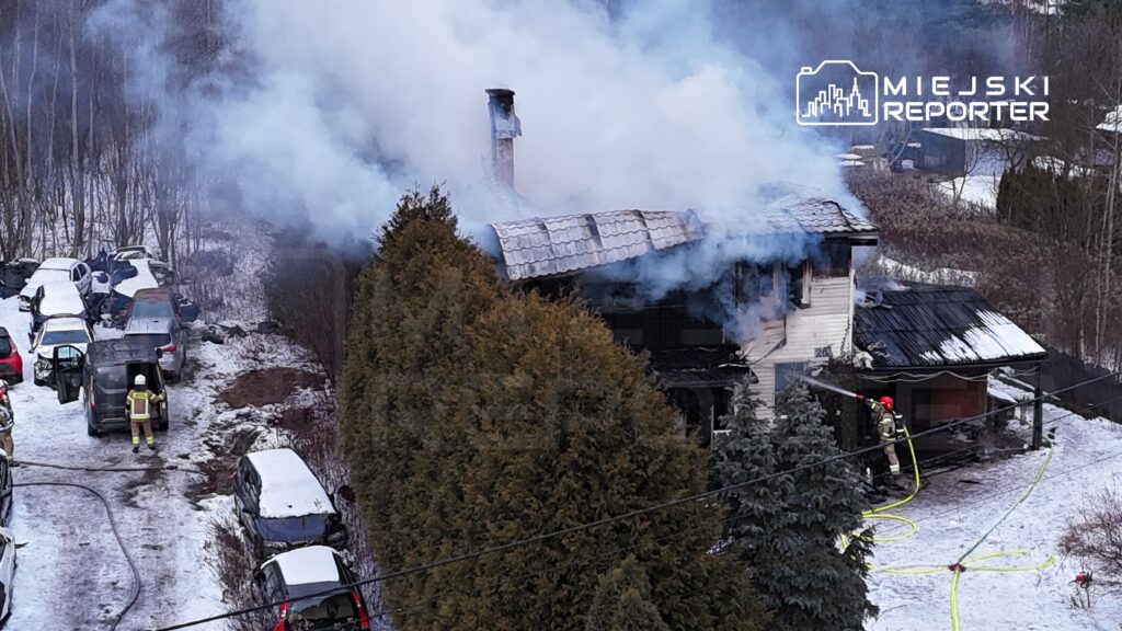 Strażak w hełmie gasi pożar dachu domu otoczonego dymem, w tle widoczne zaparkowane samochody na zaśnieżonym terenie.