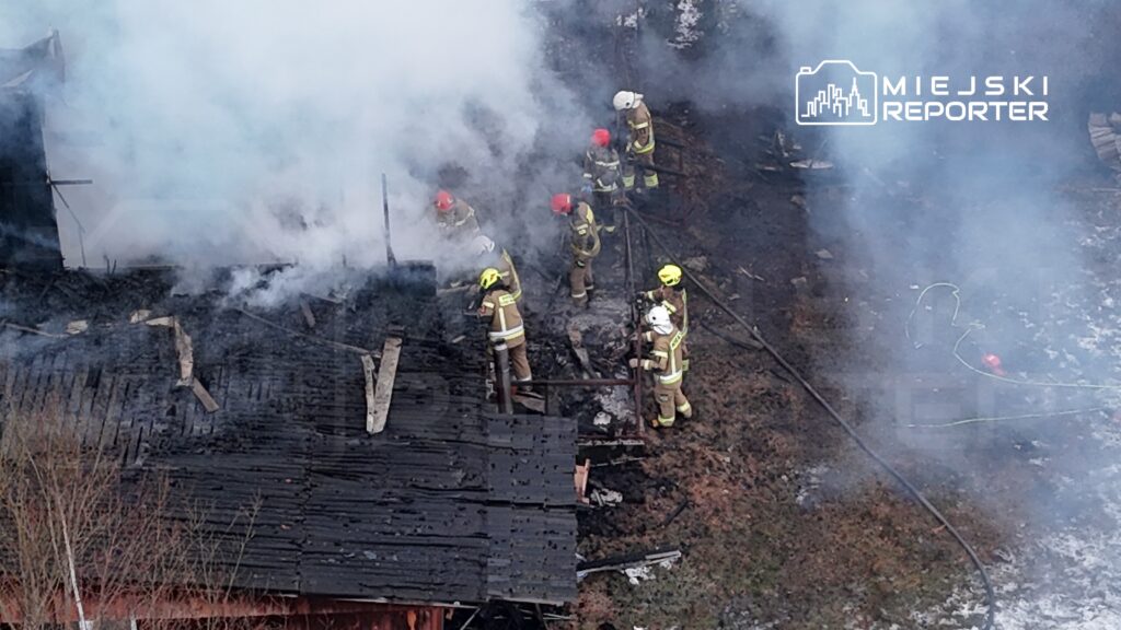 Grupa strażaków w hełmach pracuje nad ugaszeniem pożaru w zgliszczach, otoczona gęstym dymem.