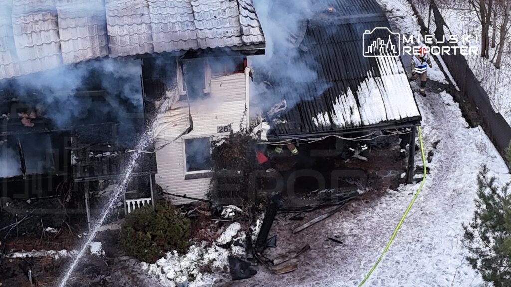 Dom z płonącym dachem, z którego wydobywa się dym, a w tle strażak gaszący ogień.