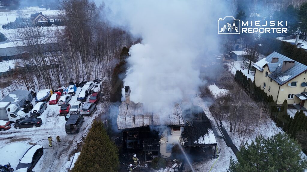 Dwa strażacy gaszą pożar w zniszczonym budynku, z którego unosi się gęsty dym, otoczonym śniegiem i zaparkowanymi samochodami.