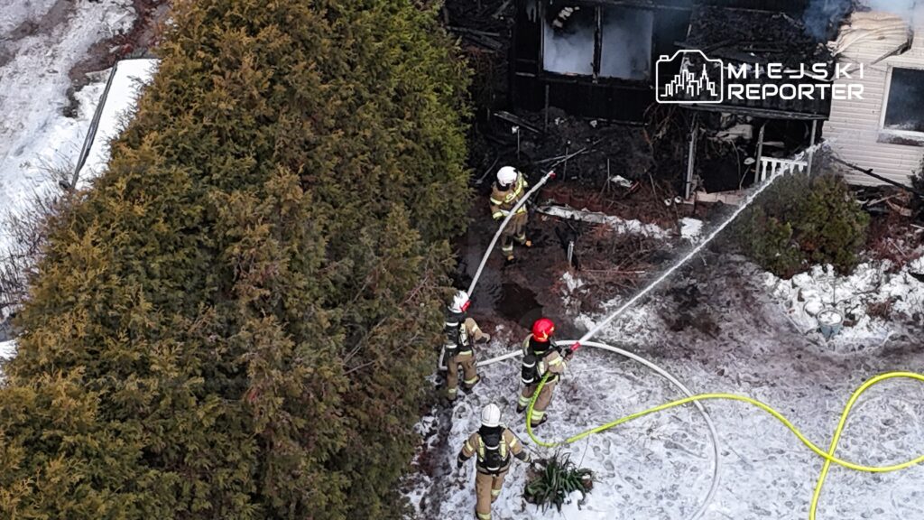 Czterech strażaków w hełmach gasi pożar zniszczonego domu, otoczonego przez gęste krzewy i śnieg.