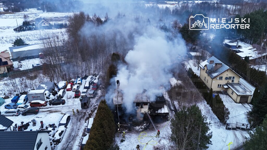 Strażacy gaszą pożar zniszczonego budynku, otoczeni przez unoszący się dym w zaśnieżonym terenie z zaparkowanymi samochodami.