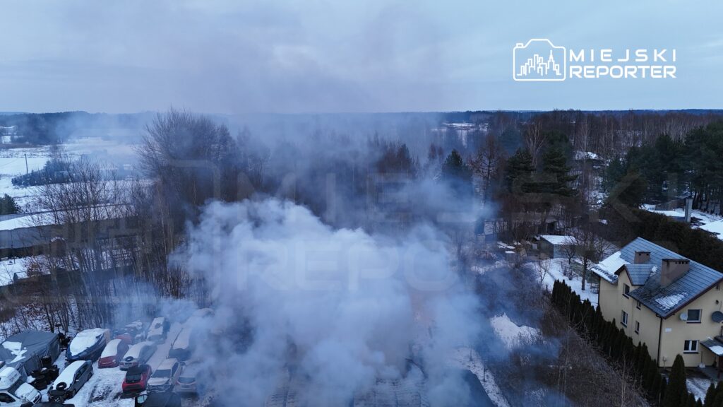 Gęsty dym unoszący się nad zadrzewionym terenem w zimowej scenerii, w tle widoczne zaparkowane samochody.