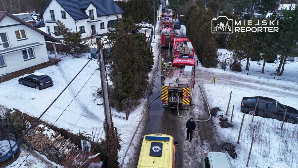Strażacy pracują przy wozach strażackich na zaśnieżonej ulicy, obok domów jednorodzinnych.