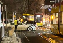 Zdarzenie drogowe na torowisku, z białym samochodem osobowym i tramwajem w tle, w nocy w mieście.