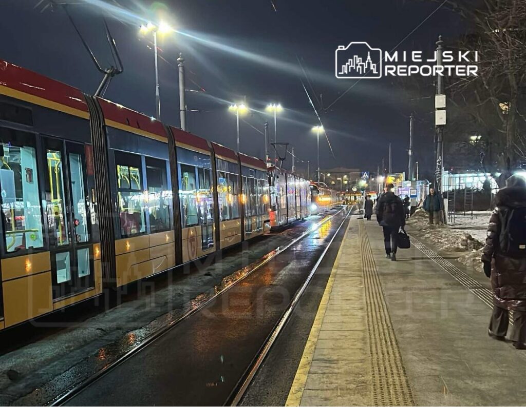 Tramwaj zatrzymany na przystanku w nocy, pasażerowie czekają na peronie oświetlonym lampami.