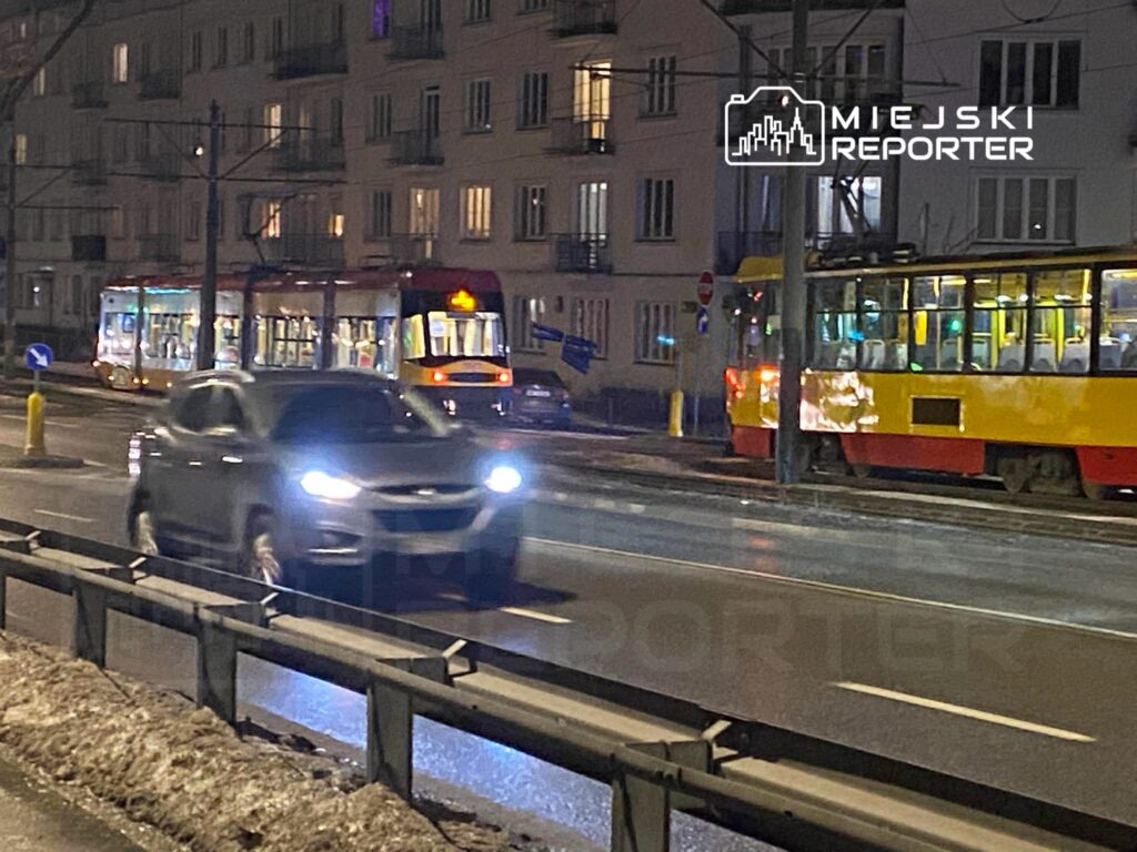 Czarny samochód osobowy przejeżdża obok tramwajów na ruchliwej ulicy w nocy, obok oświetlonych budynków mieszkalnych.