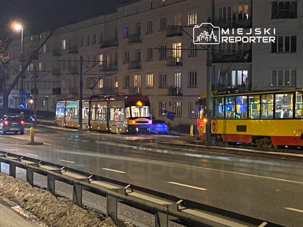 Tramwaj z podświetlonymi oknami zatrzymany na przystanku przy oświetlonej ulicy nocą. W tle widoczne budynki mieszkalne.