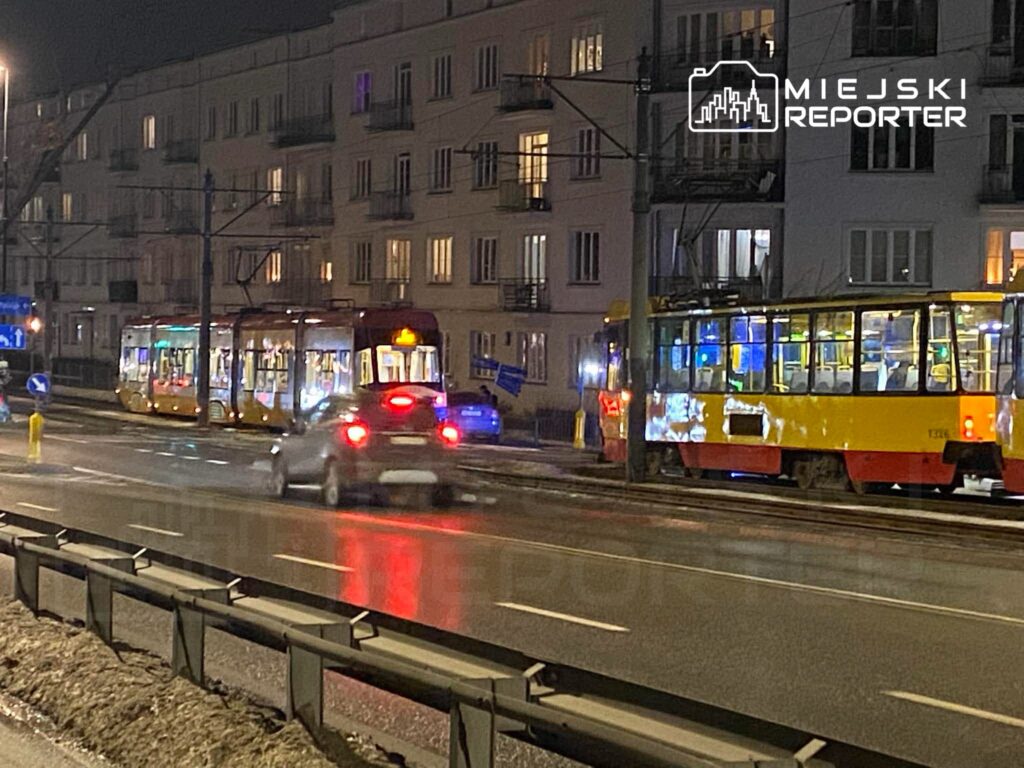 Tramwaj w nocy zatrzymany na przystanku, obok przejeżdżający samochód oraz oświetlone okna budynków mieszkalnych.