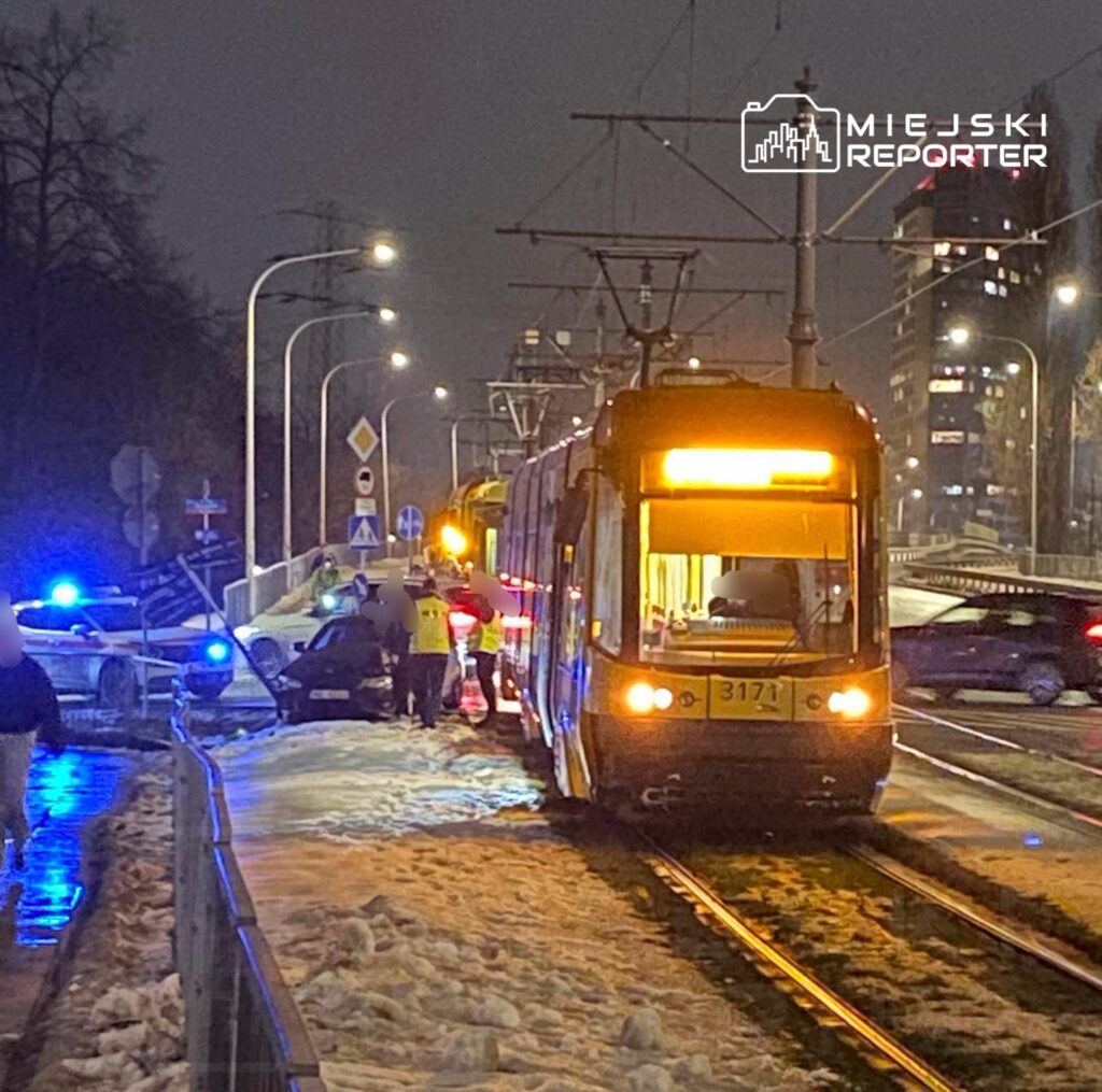 Tramwaj zatrzymany na torach, obok policjanci i ratownicy medyczni pomagający poszkodowanym przy samochodzie na zaśnieżonej jezdni.
