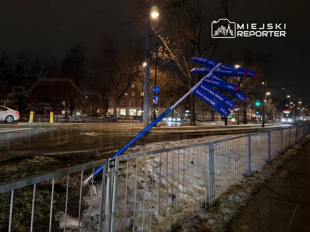Złamany słup drogowy z nieczytelnymi kierunkowskazami stoi przy oświetlonej ulicy, w tle widać tramwaj.