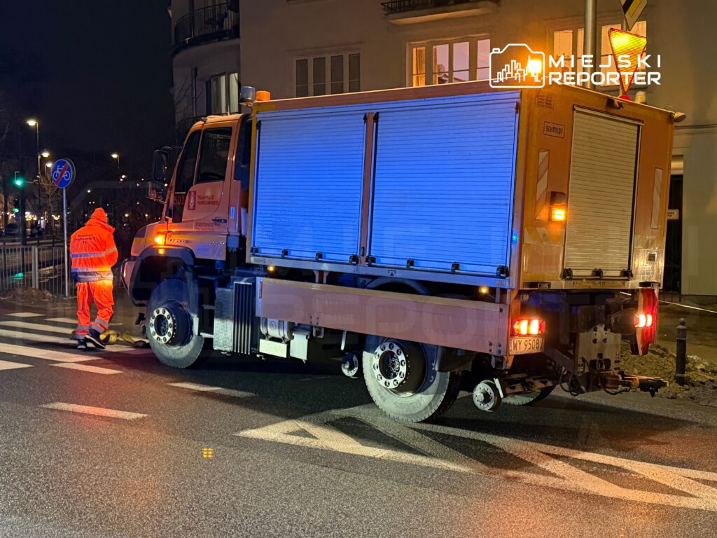Pracownik w pomarańczowym uniformie naprawia nawierzchnię drogi obok pojazdu miejskiego w nocy.