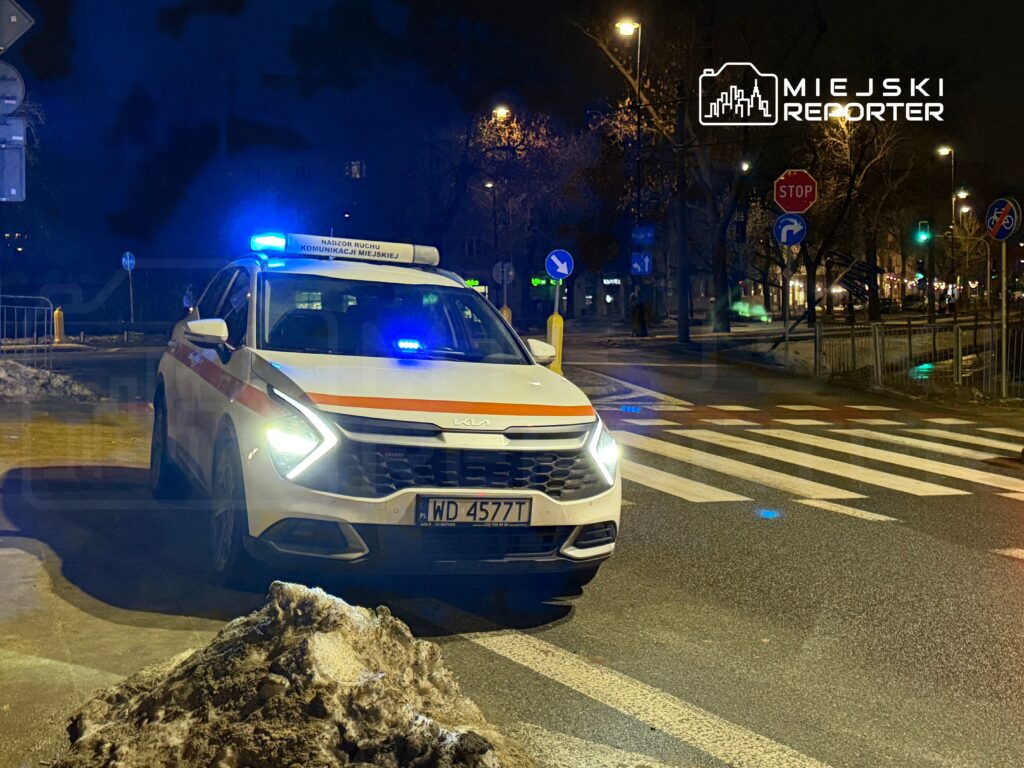 Samochód policyjny z niebieskimi światłami zatrzymany na skrzyżowaniu w nocy, obok zaspy śnieżnej.