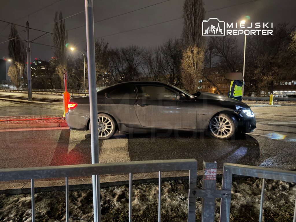 Czarny samochód osobowy zatrzymany obok torów tramwajowych, obok stoi osoba w żółtej kamizelce odblaskowej.