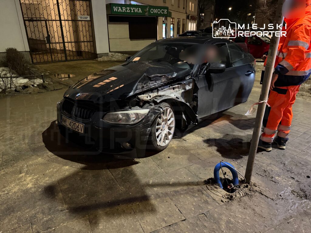Zniszczony czarny samochód osobowy zaparkowany przy chodniku, obok stoi pracownik w pomarańczowym uniformie.