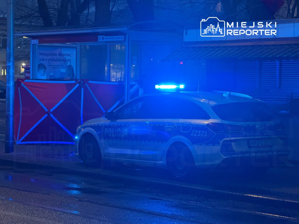 Policyjny radiowóz z niebieskimi światłami stojący obok przystanku, na którym zawieszono czerwony parawan.