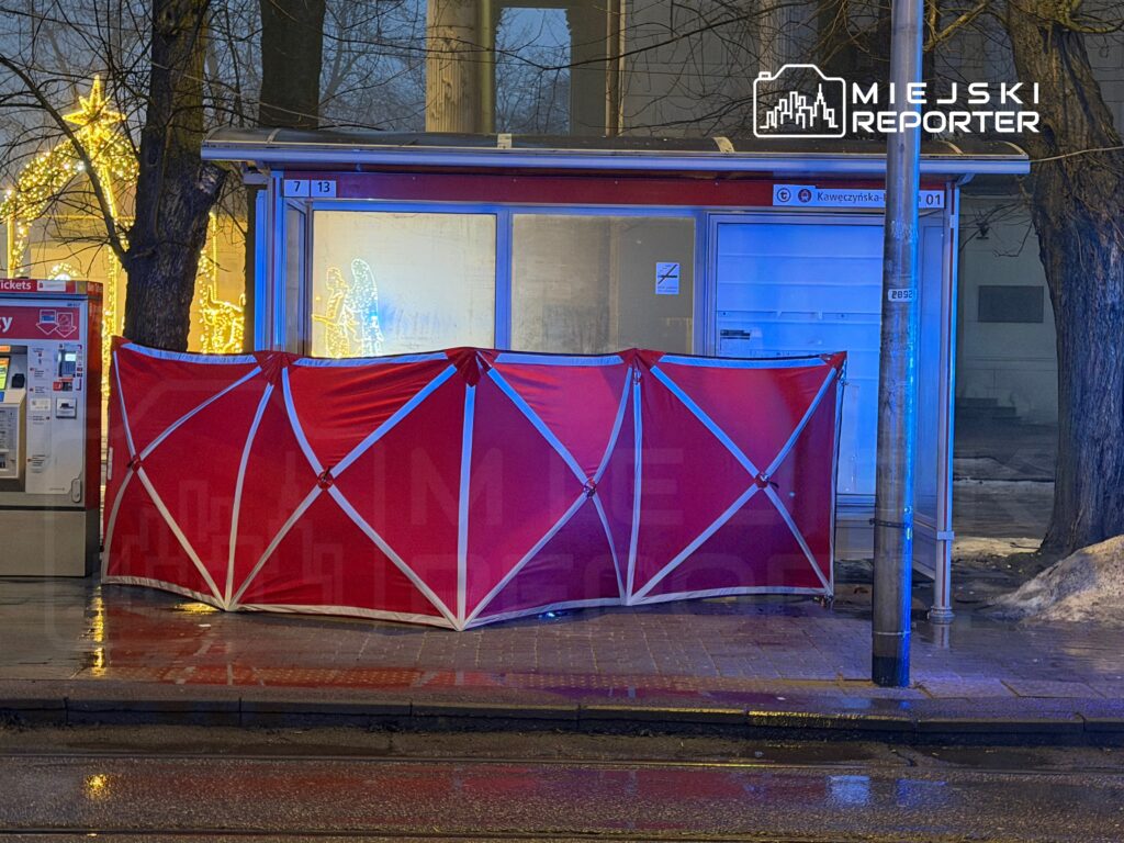 Przystanek autobusowy z czerwonym zadaszeniem, obok biletomat i świąteczne iluminacje w tle.