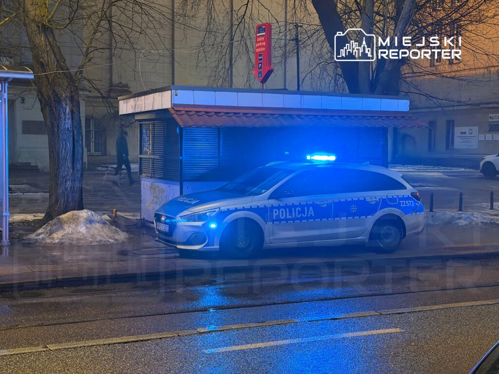Policyjny radiowóz z niebieskimi światłami zaparkowany obok kiosku w oświetlonej ulicy, w tle przechodzień.