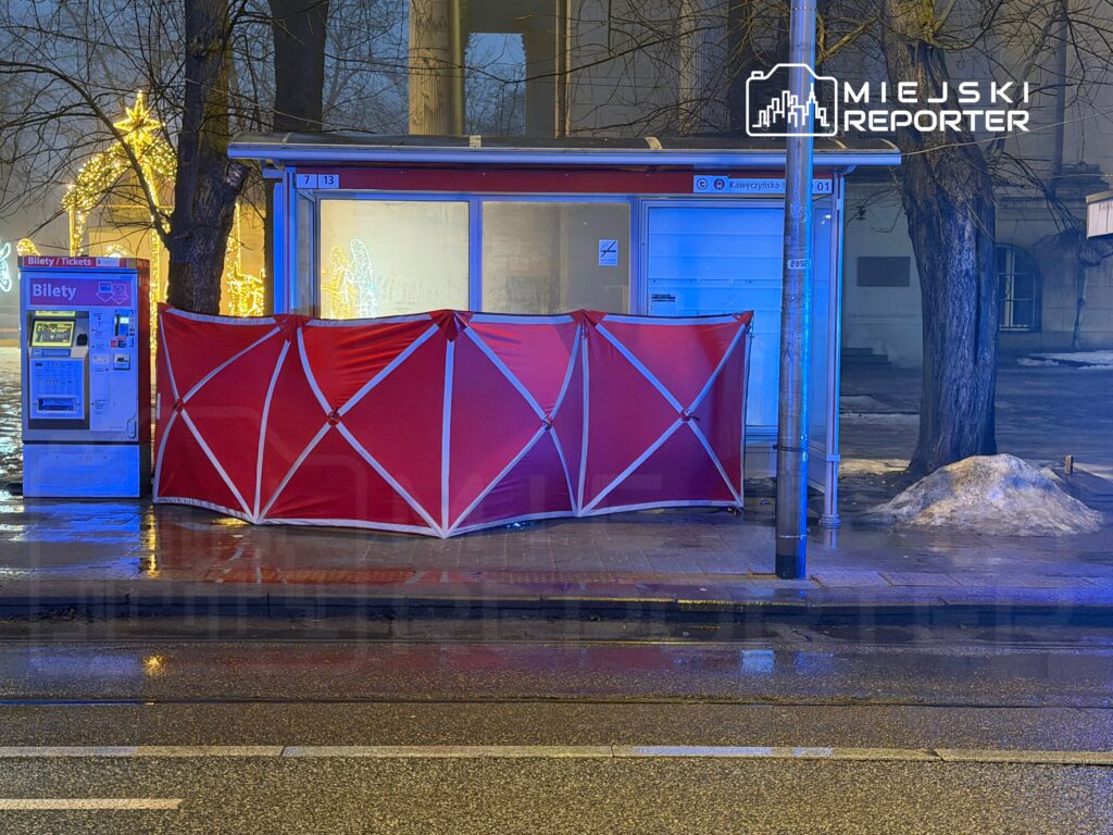 Czerwony namiot osłania kiosk biletowy przy przystanku, w otoczeniu oświetlonych dekoracji świątecznych.