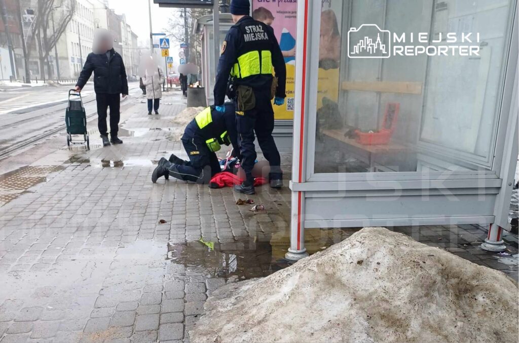 Dwaj funkcjonariusze straży miejskiej udzielają pomocy osobie leżącej na chodniku w miejskim otoczeniu.
