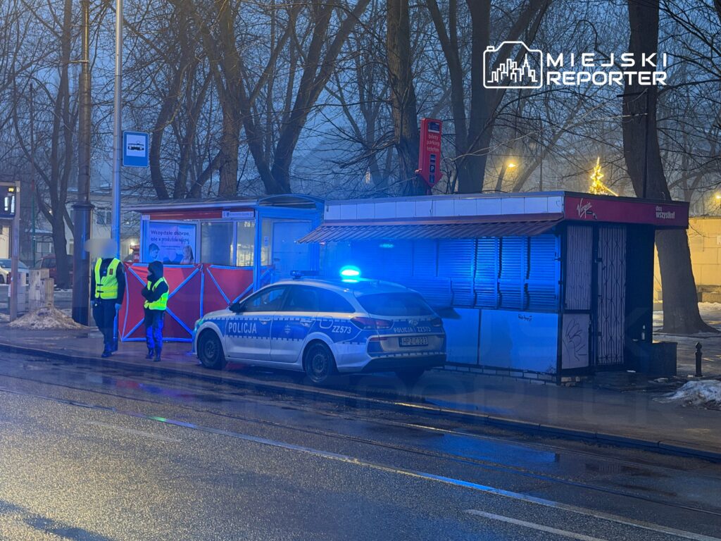 Policjanci w odblaskowych kamizelkach rozmawiają obok radiowozu z włączonymi sygnałami świetlnymi przy przystanku.