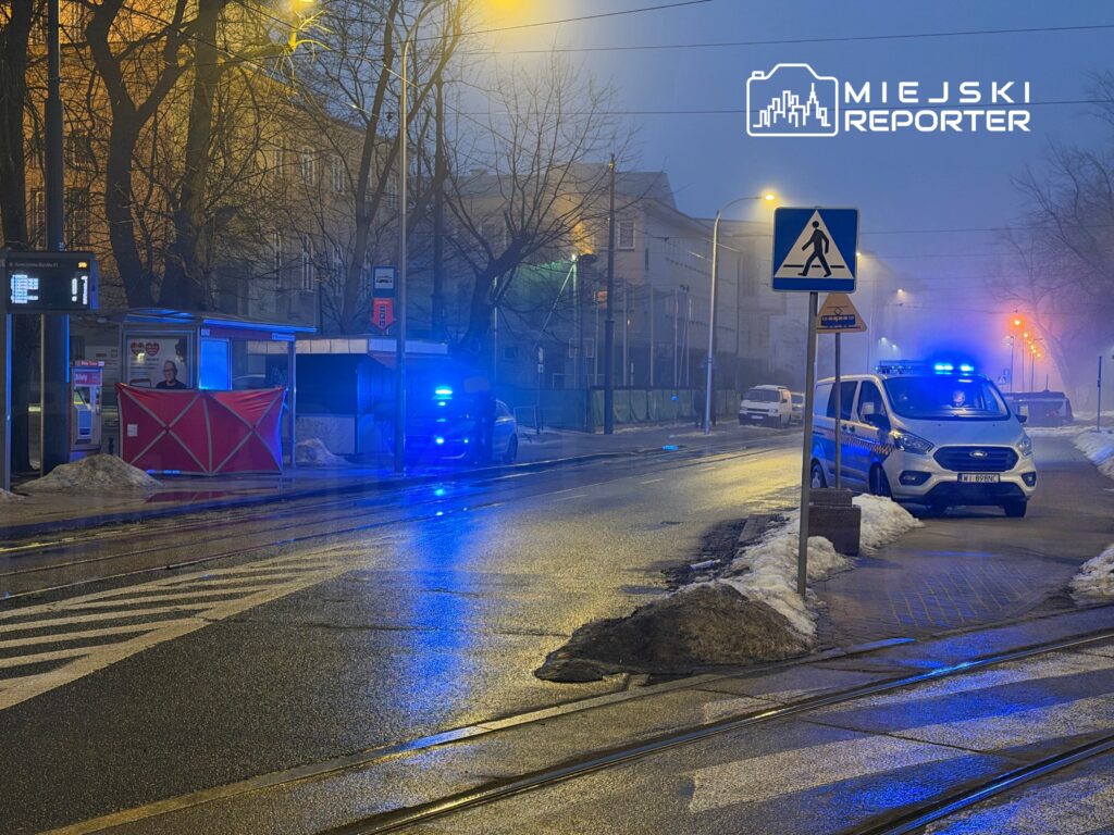 Policyjny radiowóz z niebieskimi światłami zatrzymany na ulicy w mglisty wieczór, obok przystanku z budką.