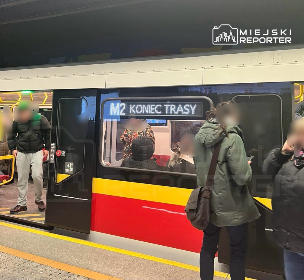 Pasażerowie wysiadają z metra na stacji, obok wyświetlacza z napisem „M2 KONIEC TRASY”.