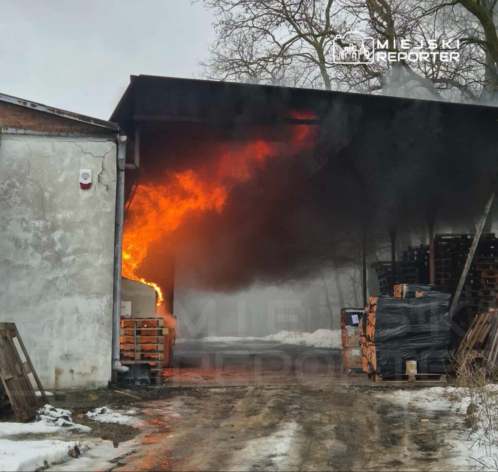 Płomienie wydobywające się z budynku, otoczone gęstym dymem, w pobliżu stosów drewnianych palet.