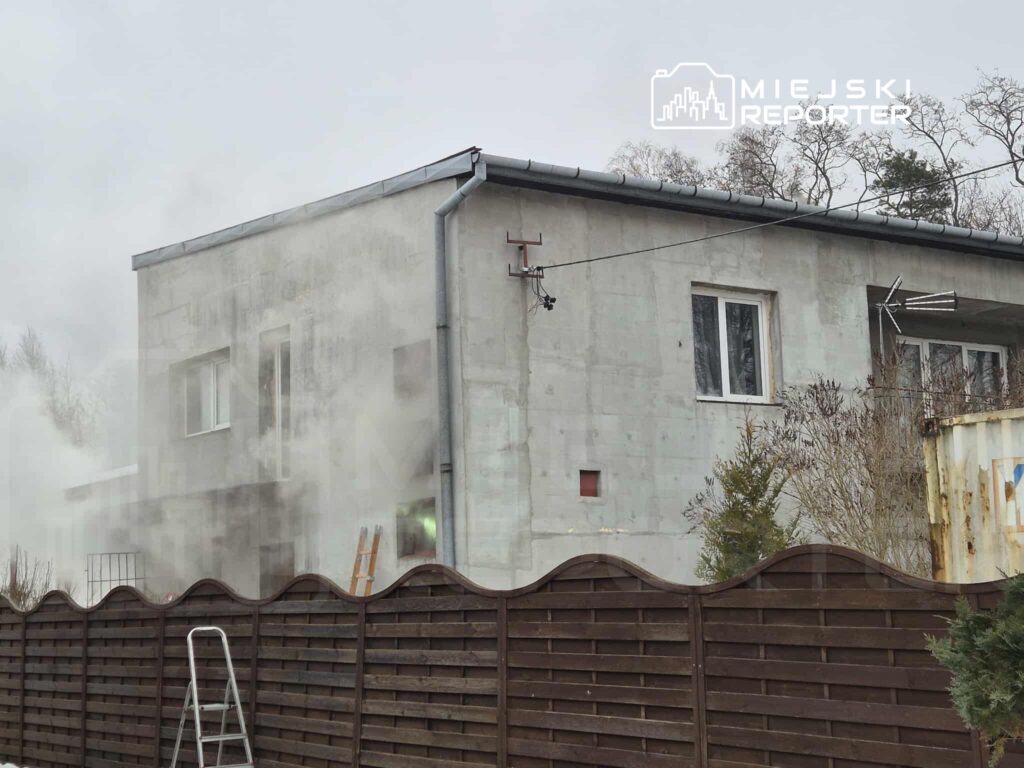 Dom jednorodzinny z surowym betonowym wykończeniem, w tle unoszący się dym, w otoczeniu ogrodzenia i drzew.