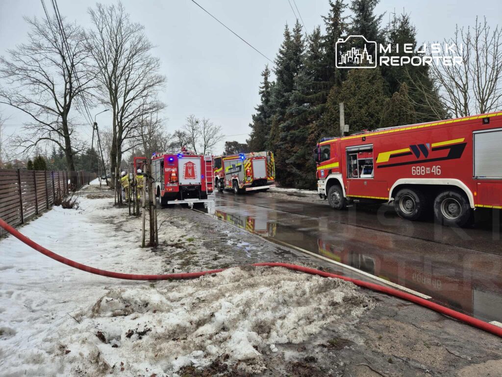 Cztery wozy strażackie na drodze, obok leży wąż strażacki, a w tle widać drzewa i resztki śniegu.