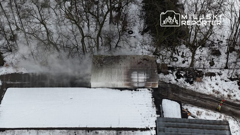 Dym wydobywający się z dachu zniszczonego budynku w lesie, w otoczeniu śniegu i drzew, obok przechodzą strażacy.