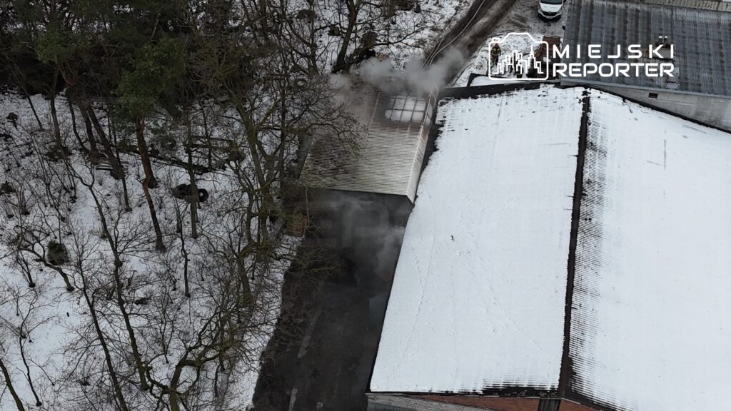 Dym wydobywający się z dachu budynku w otoczeniu zaśnieżonego terenu i drzew.