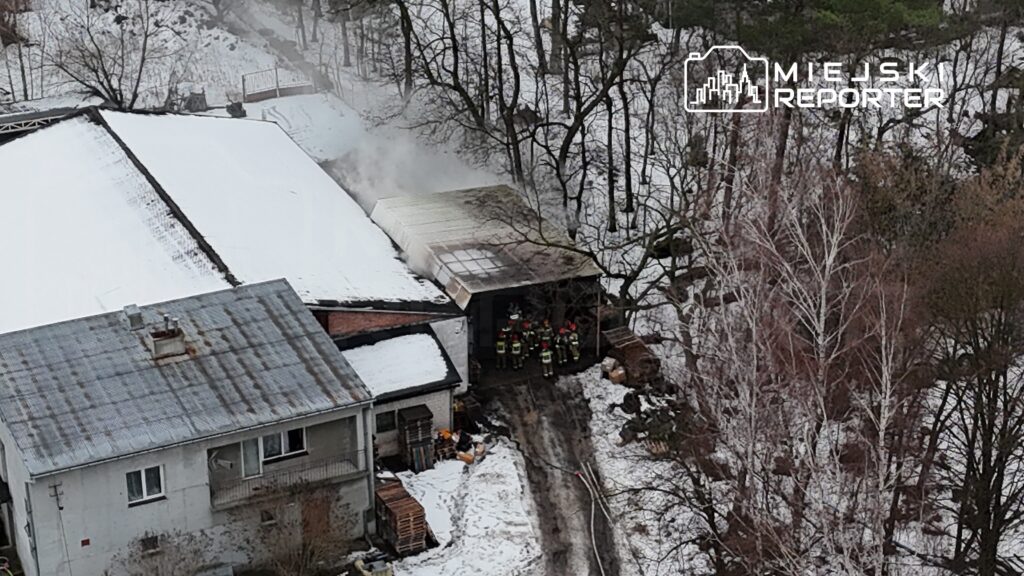Grupa strażaków w hełmach i mundurach przygotowuje się do akcji gaśniczej w pobliżu palącego się budynku.