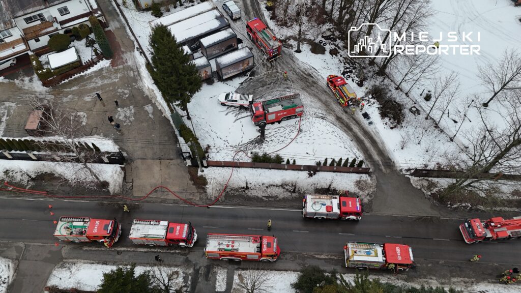Cztery wozy strażackie na zaśnieżonej ulicy, strażacy pracują przy usuwaniu skutków zdarzenia w okolicy budynków mieszkalnych.