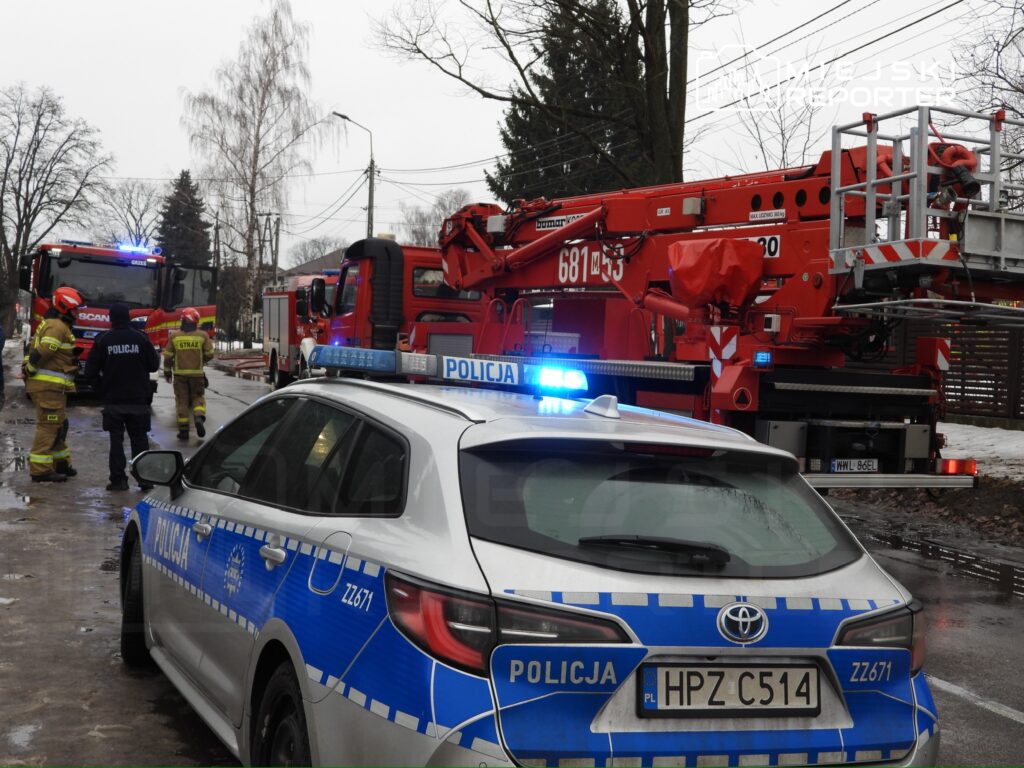 Grupa strażaków i policjantów pracuje przy wozach strażackich na ulicy, obok podnośnika strażackiego.