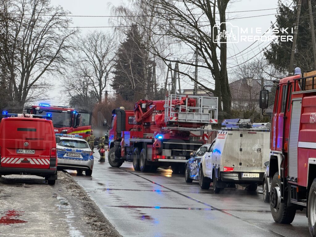 Na ulicy zaparkowane pojazdy straży pożarnej i policji, w tle widać strażaków pracujących przy akcji ratunkowej.