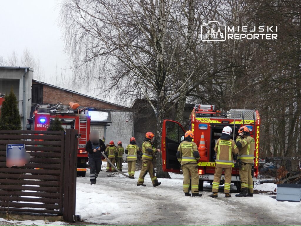 Grupa strażaków w mundurach z hełmami przygotowuje sprzęt przy wozie strażackim na zaśnieżonej drodze.
