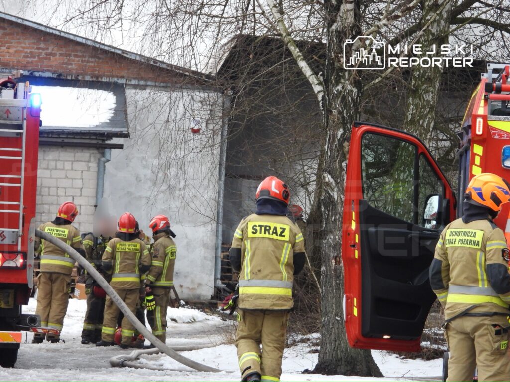 Grupa strażaków w mundurach omawia działania ratunkowe obok wozu strażackiego przed budynkiem z dymem.