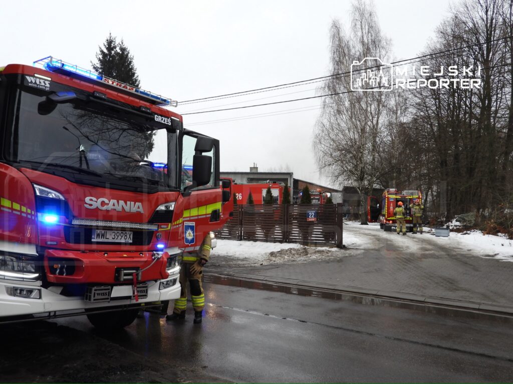 Czerwony wóz strażacki z włączonymi sygnałami świetlnymi stoi na drodze, obok grupy strażaków działających w zimowym otoczeniu.