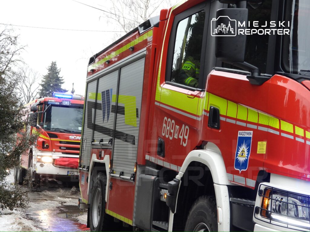 Czerwony wóz strażacki z włączonymi sygnalizatorami na drodze, obok drugi wóz strażacki w tle.