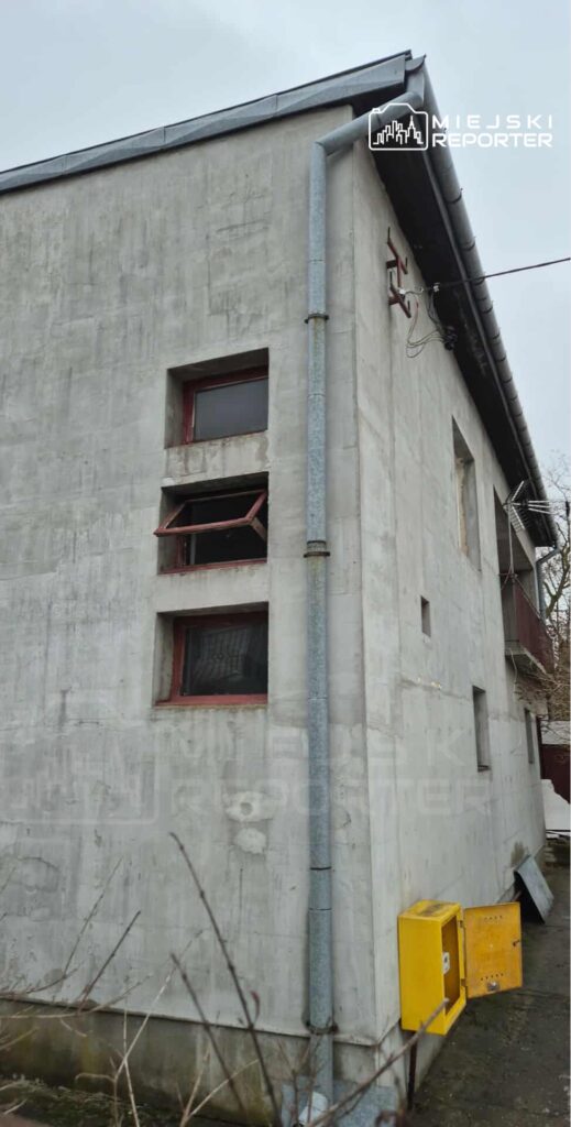 Szary budynek z tynkiem, z otwartymi oknami i metalowym odpływem, obok żółta skrzynka na urządzenia.