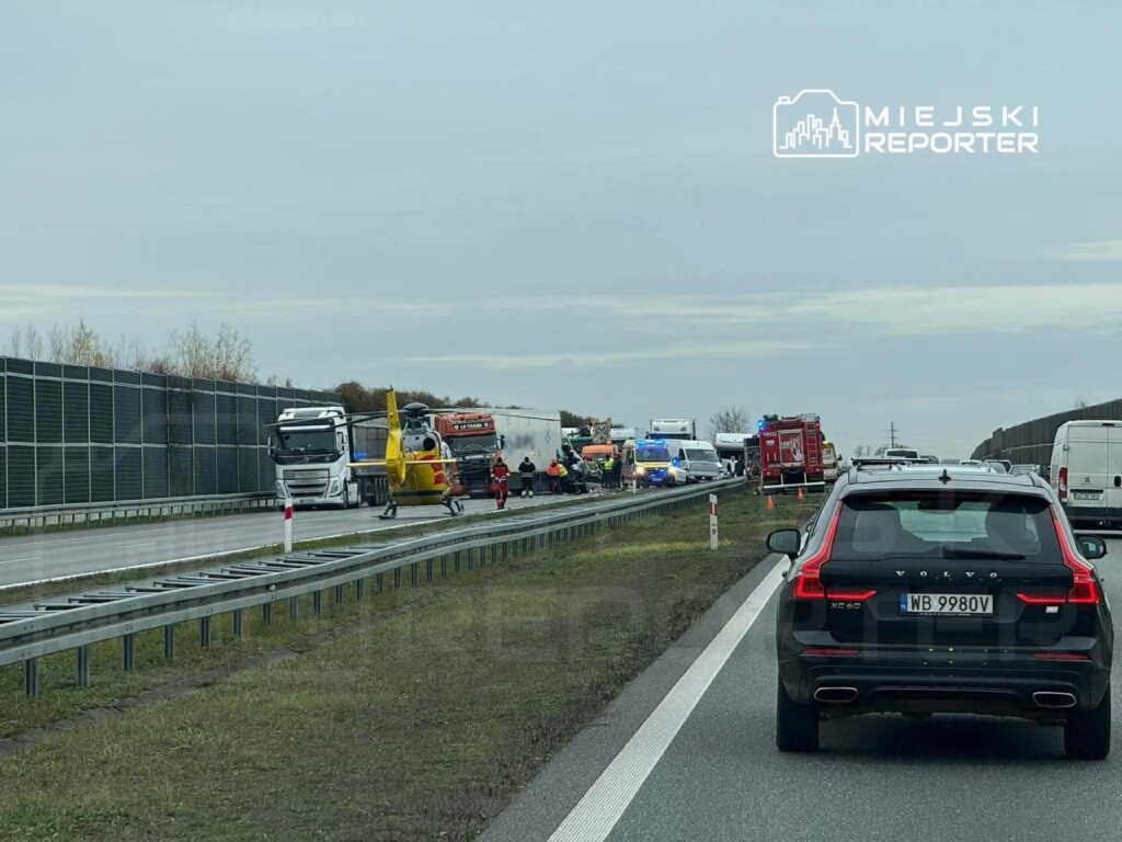 Na drodze stoi czarny samochód osobowy, obok widać karetki, straż pożarną oraz helikopter ratunkowy. W tle zablokowane ciężarówki.