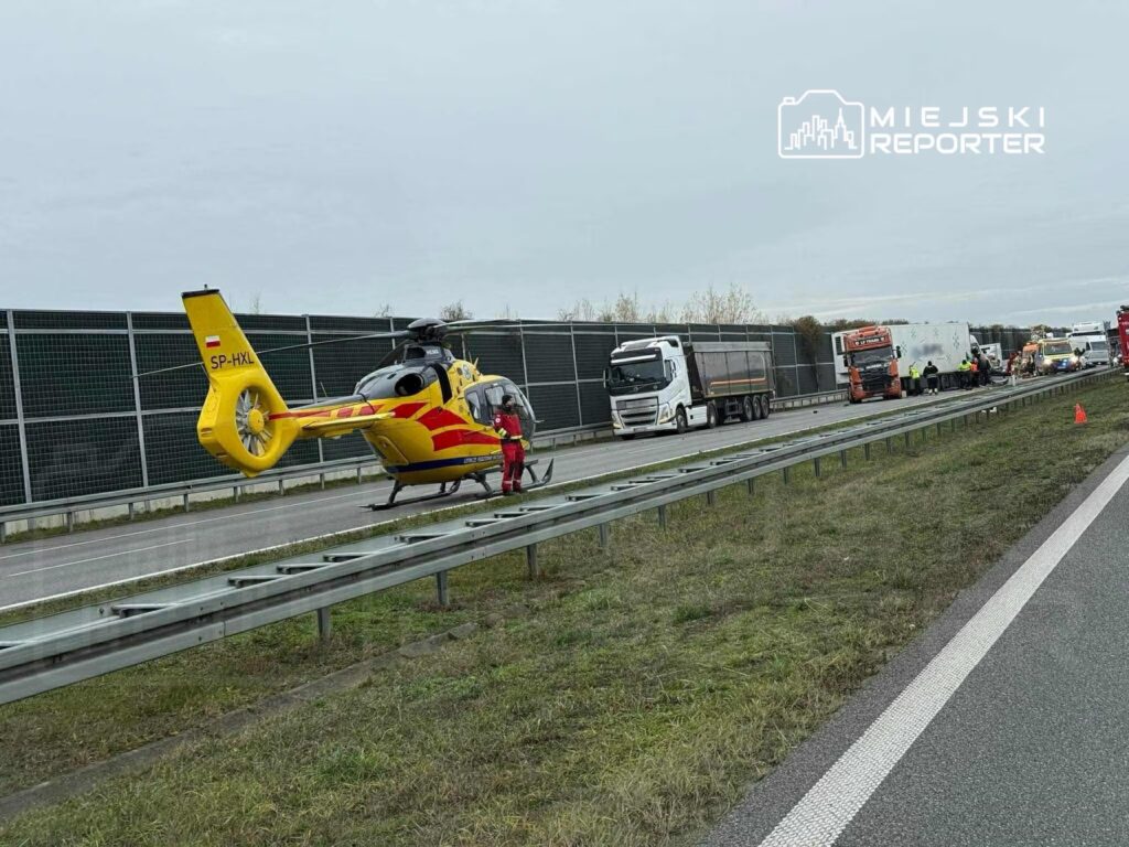 Żółty helikopter ratunkowy ląduje obok zatoru drogowego na autostradzie, obok ciężarówek i pojazdów służb.